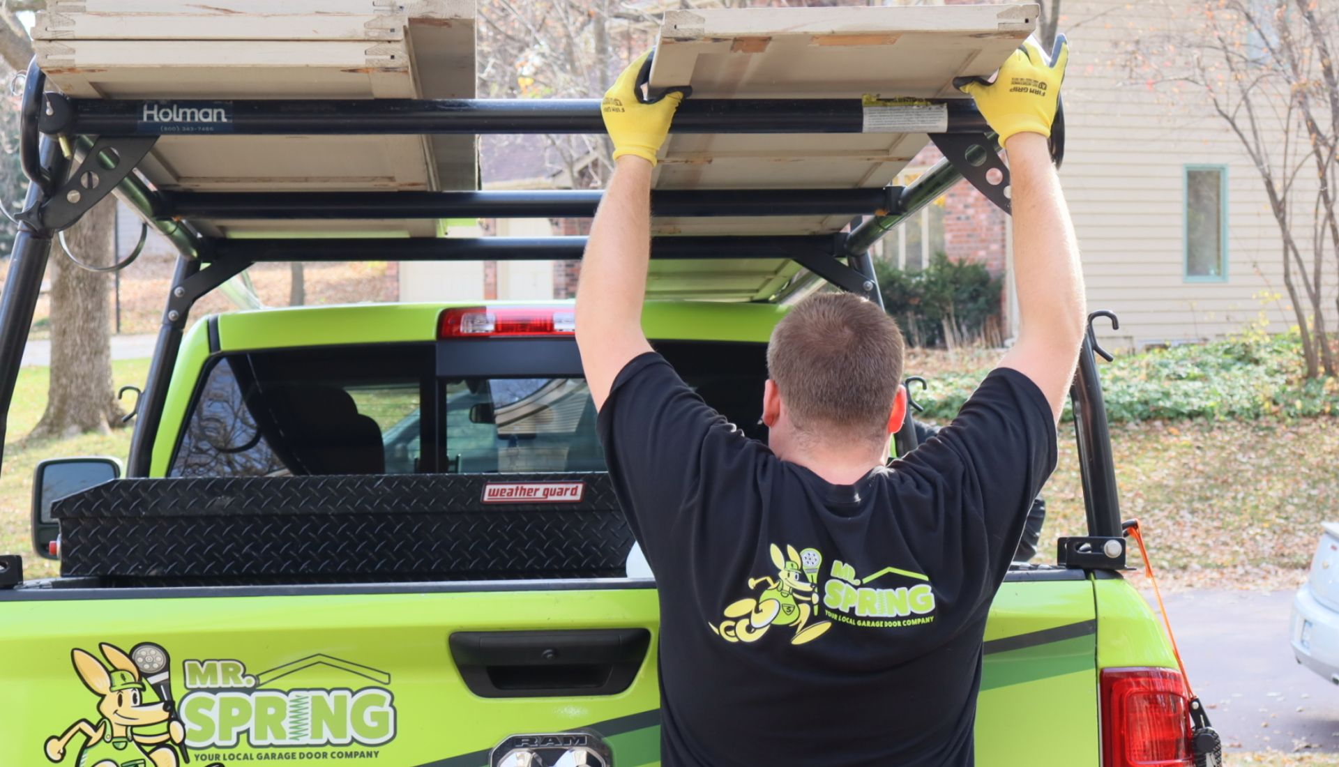 A repair tech reaches overhead to remove a garage door panel from the rack on a bright green Mr. Spring truck