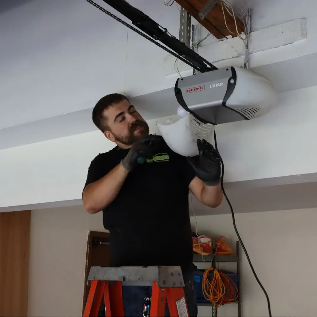 repair technician wearing gloves uses a ladder to reach and remove the cover of a garage door opener light